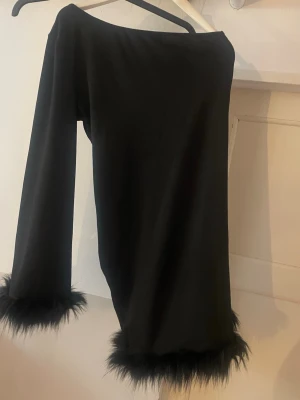 Svart klänning med fuskpälsdetaljer - Stilren svart klänning med långa ärmar och trendiga fuskpälsdetaljer vid ärmslut och nederkant. Klänningen har en rak passform och är perfekt för dig som gillar en edgy och unik look.