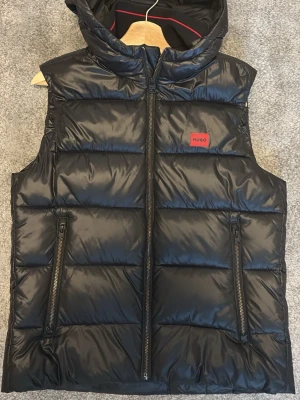 Svart quiltad dunväst från Hugo - Snygg svart dunväst från Hugo med quiltad design och huva. Västen har två dragkedjeförsedda fickor framtill och en röd Hugo-logga på bröstet. Perfekt för lager på lager och streetwear-vibe.