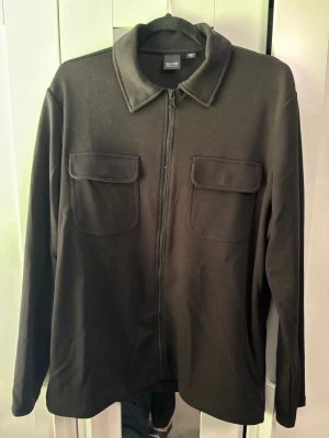 Svart overshirt med dragkedja - Stilren svart overshirt från H&M med klassisk krage och två bröstfickor med lock. Jackan har dragkedja framtill och är tillverkad i ett mjukt, strukturerat material. Perfekt att slänga över en t-shirt för en clean look.