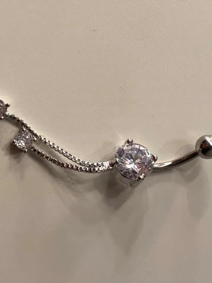 Snygg navelpiercing med strass - Unik navelpiercing i silverfärgad metall med en stor rund klar sten och mindre gnistrande stenar på kedjor. Elegant design med två tunna kedjor som ger extra detalj. Helt oanvänd och i rostfritt stål! 🫶