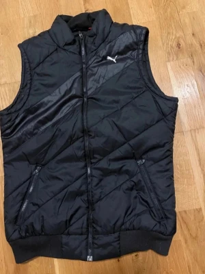 Svart väst från Puma stl XSmall  - Snygg svart väst från Puma med quiltad design och dragkedja framtill. Västen har två fickor med dragkedja och en hög krage. Perfekt för lager-på-lager och streetstyle.