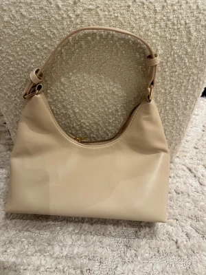 Beige axelremsväska i skinnimitation - Stilren beige handväska med axelrem och guldfärgade metalldetaljer. Väskan har en mjuk, något rundad form och stängs med dragkedja upptill. Tillverkad i skinnimitation och passar perfekt till en trendig vardagslook.