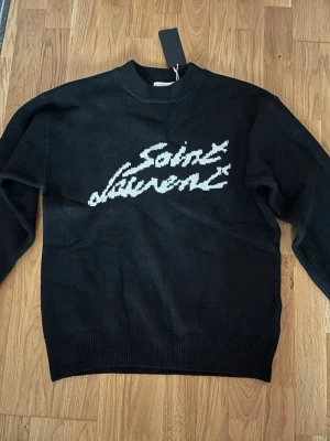 Svart Saint Laurent stickad tröja - Svart stickad tröja från Saint Laurent med vit logotyp i handskriven stil på bröstet. Tröjan har rund halsringning, ribbade muddar och är tillverkad i ett mjukt material som känns lyxigt. Perfekt för dig som gillar exklusiva och stilrena plagg.