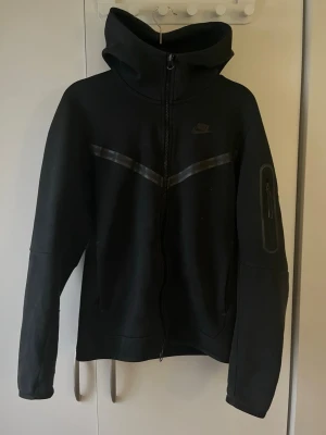 Svart Nike Tech Fleece hoodie - Säljer en svart Nike Tech Fleece hoodie med dragkedja framtill och stor huva. Perfekt för dig som gillar streetwear och vill ha något stilrent och bekvämt. Jag säljer den eftersom jag inte tycker den längre. Storlek S, pris diskuteras !