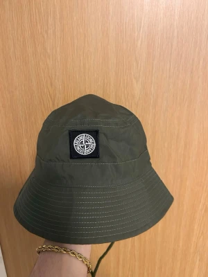 Grön bucket hat från Stone Island - Snygg grön bucket hat från Stone Island med klassisk kompasslogga framtill. Hatten är tillverkad i lätt nylonmaterial och har justerbar dragsko under hakan. Perfekt accessoar för en streetwear-look.