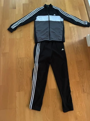 Svart och grå Adidas träningsset - Säljer ett snyggt träningsset från Adidas med svart och grå zip-jacka och matchande svarta byxor. Setet har de klassiska vita Adidas-ränderna längs ärmar och ben samt Adidas-logga på både jacka och byxa. Perfekt för träning eller chill. Materialet är mjukt och stretchigt.