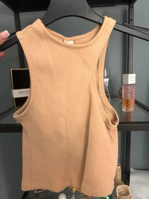 Beige ribbad linnetopp - Snygg beige ribbad linnetopp med hög halsringning och ärmlös design. Toppen har en croppad passform och är tillverkad i mjukt bomullsmaterial, perfekt för en trendig och avslappnad stil.