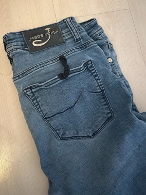 Blå jeans från Jacob Cohen - Snygga blå jeans från Jacob Cohen med klassisk femficksmodell och diskreta kontrastsömmar. Jeansen har en stilren look och är tillverkade i mjukt denimtyg som ger en bekväm passform. Perfekta för dig som gillar tidlös och avslappnad stil. Små defekter på båda bakfickorna samt nere på vänster benet och en under gylfen