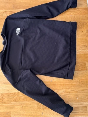 Svart tröja från The North Face - Svart långärmad tröja från The North Face med vit logga på bröstet. Tröjan har en sportig look, rund halsringning och är tillverkad i ett strukturerat, tekniskt material som känns lätt och bekvämt.