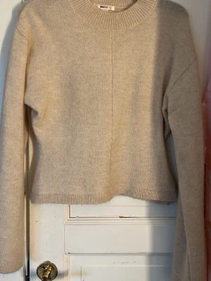 Beige stickad tröja - En enkel och stilren beige stickad tröja med rund halsringning och långa ärmar. Tröjan har en rak passform och ribbade muddar vid ärmslut och nederkant. Perfekt basplagg till garderoben och lätt att matcha med olika outfits.