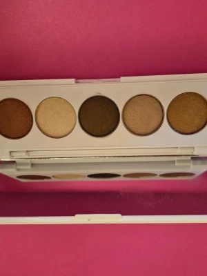 Ögonskuggspalett från OFRA i neutrala toner - Säljer en ögonskuggspalett från OFRA med fem runda skuggor i neutrala nyanser: koppar, champagne, mörkbrun, beige och guld. Paletten har ett vitt plastfodral med spegel på insidan, perfekt för sminkning on-the-go. Kan vara testad ,swatshad någon gång. Fin ögonskuggspalett.