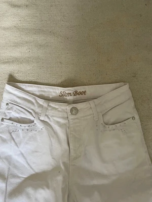 Vita bootcut jeans - Snygga vita slim bootcut jeans med dekorativa nitar på fram- och bakfickorna. Klassisk femficksmodell med bälteshällor och knappgylf. Perfekta för dig som vill ha en clean look med lite extra detaljer. Innerbensländ: 72cm, midjemått: 36cm