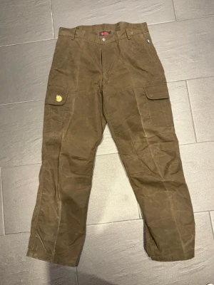 Bruna Vintage cargobyxor från Fjällräven - Snygga bruna Vintage cargobyxor från Fjällräven knappt använda, med klassisk passform och flera praktiska fickor på sidorna. Tillverkade i slitstarkt vaxat material som tål tuffa tag. Ikonisk logga på benfickan och justerbara benslut för perfekt fit. Nypris 4000kr, Storlek 50. Säljer de för 1500kr. 