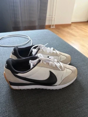 Nike vita och beige sneakers - Snygga sneakers från Nike i vitt och beige med svart swoosh på sidorna. Skorna har klassisk låg modell, vita skosnören och svart innerfoder. Ovandelen är i en mix av läder och mocka, med gummisula i vitt och brunt. Perfekta för en stilren och sportig look.