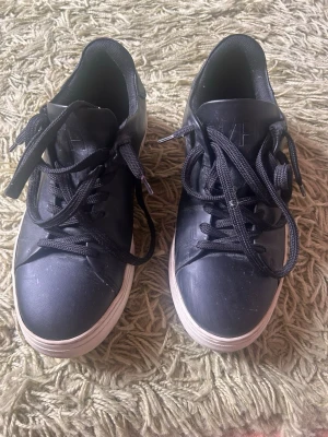 Svarta sneakers från Selected Homme - Svarta sneakers från Selected Homme med klassisk låg profil och vita sulor. Skorna har svarta skosnören och ovandel i syntetmaterial. Perfekta för en stilren och enkel look.