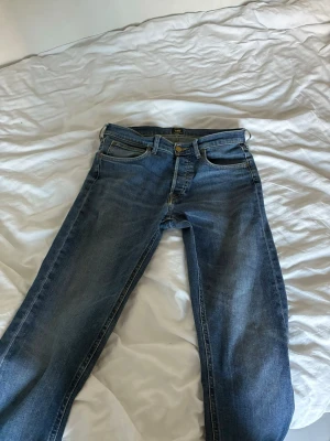 Blå Lee jeans med rak passform - Ett par klassiska blå jeans från Lee med rak passform och fem fickor. Jeansen har Lee-logga på knappen och är gjorda i slitstarkt denimtyg. Perfekta för dig som gillar enkel och tidlös stil.