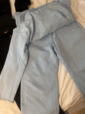 Nike NOCTA ljusblå mjukisbyxor - Säljer ett par ljusblå mjukisbyxor från Nike NOCTA-kollektionen. Byxorna har en clean look med diskret NOCTA-tryck på benet och Nike Swoosh på låret. Tillverkade i mjukt material med raka ben och elastisk midja för skön passform.