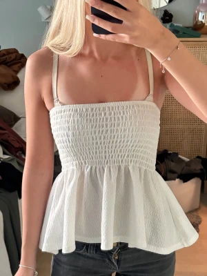 Vit smockad linnetopp från Gina Tricot - Supersöt vit linnetopp från Gina Tricot med smockad överdel och volangig nederdel. Tunt, luftigt material och smala axelband gör den perfekt som strand topp! 💕inga tecken på användning!