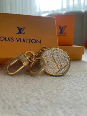  Vuitton guldfärgad nyckelring - Exklusiv nyckelring från  Vuitton i guldfärgad metall med stor rund berlock täckt av gnistrande stenar och LV-logga. Snygg detalj till väskan eller nycklarna, med både ring och karbinhake för enkel fastsättning.