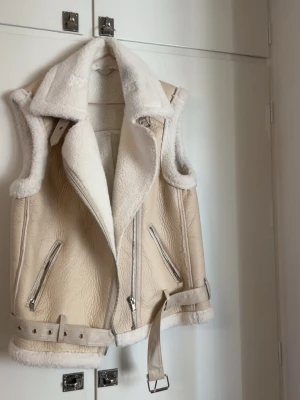 Beige pälsväst med bälte och dragkedjor - Stilren beige pälsväst från SHEIN med vit fuskpäls på insidan och krage. Västen har två snedställda dragkedjefickor, bred krage och bälte i midjan för extra detalj . Perfekt för  jeans eller kjol.