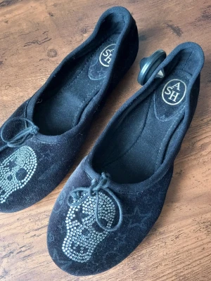 Svarta ballerinaskor från ASH med döskalle storlek 36 - Svarta ballerinaskor från ASH i sammet med coola döskallemotiv av glittrande stenar framtill. Skorna har rund tå, liten rosett och diskret stjärnmönster i materialet. Perfekta för dig som gillar edgy detaljer och vill sticka ut.