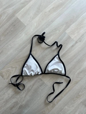 Vit och svart trekantsbikinitopp - Säljer en stilren trekantsbikinitopp i vitt med svarta kanter och knytband i nacken och ryggen. Klassisk modell som ger en snygg siluett och passar perfekt till sommaren. Materialet känns mjukt och följsamt.