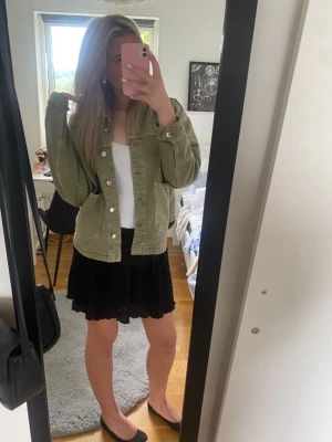 Olivgrön jeansjacka med silverknappar - Snygg olivgrön jeansjacka med klassisk krage och stora fickor framtill. Jackan har silverfärgade knappar och en avslappnad passform som ger en cool vibe. Perfekt att slänga över en t-shirt eller klänning för en trendig look.