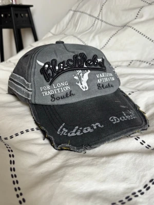 Grå keps BlackRebel med tryck - Snygg grå keps från BlackRebel med broderad text och tryck framtill. Kepsen har slitna detaljer, böjd skärm och texten 'Indian Dakota' på skärmen. Materialet är bomull och kepsen har justerbar passform baktill.