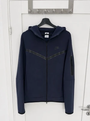 Mörkblå Nike Tech Fleece hoodie - Mörkblå Nike Tech Fleece hoodie med hel dragkedja och huva. Har en diskret Nike-logga på bröstet och en svart detalj över bröstet. Praktisk ficka med dragkedja på ärmen och två sidofickor. 
