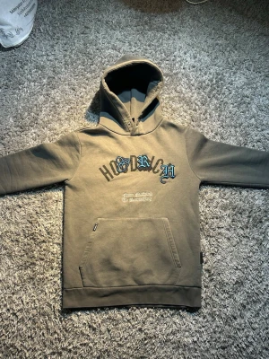  hoodie från Hoodrich - Snygg  hoodie från Hoodrich med tryck i svart och blått framtill och stort tryck på ryggen. Klassisk känguruficka och huva. Perfekt för en streetwear-look och chill vardagsstil.