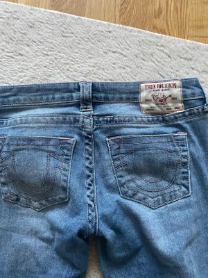 True Religion ljusblå jeans - Säljer ett par lågmidjade ljusblå jeans från True Religion med klassiska bakfickor och kontrastsömmar. Jeansen har en patch med logga och coola detaljer som sticker ut. Dem är flare.