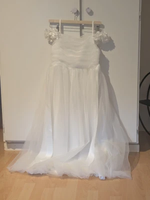 Vit långklänning - Vit långklänning med axelband dekorerade med blommor. Klänningen har en luftig kjol i flera lager tyll och en enkel, elegant överdel. Perfekt för speciella tillfällen.