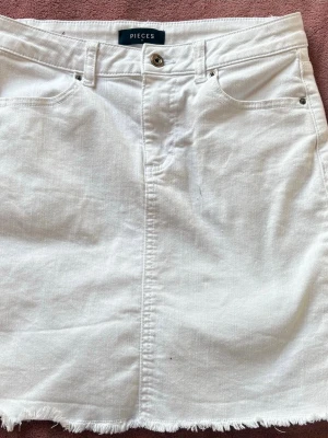 Vit jeanskjol från Pieces - Vit kort jeanskjol från Pieces med rå kant nertill, klassiska fem fickor och bälteshällor. Kjolen har knapp och dragkedja framtill och är perfekt för en trendig och avslappnad look.