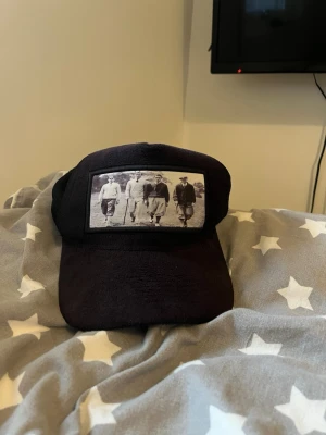 Emilio pendenza velvet keps - Säljer en svart velvet keps med mesh på sidorna och ett stort vintage-inspirerat tryck framtill som föreställer fyra personer i svartvitt. Kepsen har böjd skärm och justerbar baksida för skön passform. Perfekt för dig som gillar retrostil och streetwear.