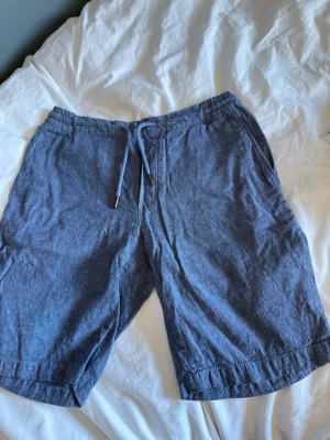 Blå linneshorts från Dressmann - Säljer ett par blå linneshorts från Dressmann i storlek S. Shortsen har elastisk midja med snörning och två sidofickor. Perfekta för varma dagar och har en avslappnad passform. Materialet är svalt och skönt linne.