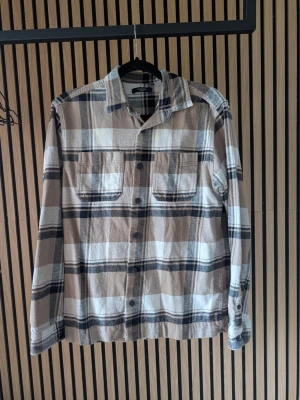 Rutig overshirt från Jack & Jones - Snygg overshirt i beige, brunt och svart rutmönster från Jack & Jones. Skjortan har två bröstfickor med lock, knäppning framtill och klassisk krage. Perfekt lager-på-lager-plagg i mjukt bomullstyg.