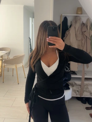 Svart omlottopp med knyt - Svart omlottopp med knyt i sidan och v-ringad hals. Toppen har långa ärmar och sitter tight. Under syns en vit topp med djup v-ringning. Perfekt för en trendig och stilren look.