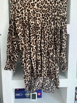Leopardmönstrad klänning från H&M - Säljer en långärmad klänning från H&M med leopardmönster i beige och svart. Klänningen har lös passform och är tillverkad i ett mjukt material som känns skönt mot huden. Perfekt för dig som vill sticka ut med ett djurmönstrat plagg.