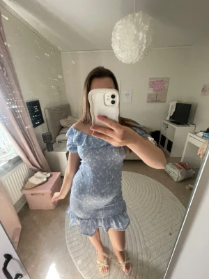 Blå offshoulder klänning med volang - Söt blå kortklänning med vita blommor, offshoulder-modell och volang nertill. Klänningen har en figurnära passform och är perfekt för sommarens varma dagar. Tillverkad i ett lätt och bekvämt material.