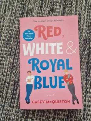 Red, White & Royal Blue bok - Ny - Säljer boken 'Red, White & Royal Blue' av Casey McQuiston. Omslaget är rosa med färgglad text i rött, vitt och blått samt illustrationer av två personer. Pocketbok i mjukt papper, perfekt för dig som gillar romantisk och modern skönlitteratur. Det finns fukt skador på toppen av sidorna, men inget man tänker på. Oläst.