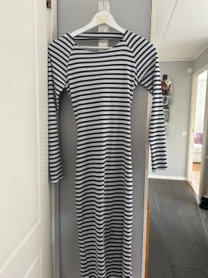 Randig långärmad långklänning 💙🤍 - Snygg långklänning med svartvita ränder och lång ärm. Klänningen har en unik omlottdetalj i ryggen som ger en cool twist. Tillverkad i mjukt ribbat material som sitter skönt mot kroppen. Perfekt för dig som gillar en stilren men ändå trendig look.