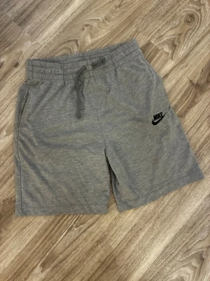 Gråa sweatshorts från Nike - Gråa mjuka sweatshorts från Nike med resårmidja och dragsko. Klassisk svart Nike-logga på vänster ben. Shortsen har sidofickor och är tillverkade i ett bekvämt bomullsmaterial, perfekta för chill eller träning. (350)