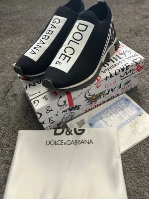 Dolce & Gabbana svarta sneakers - Svarta slip-on sneakers från Dolce & Gabbana med stora vita logotyper på ovandelen och sidorna. Skorna har en sportig design med ribbad sula och elastisk ovandel i textil för en bekväm passform. Snygga detaljer och tydlig branding gör dem till ett statement.