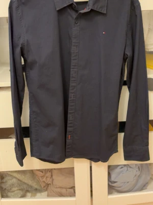 Mörkblå skjorta från Tommy Hilfiger - Stilren svart skjorta från Tommy Hilfiger med klassisk krage och diskret logga på bröstet. Skjortan har långa ärmar och knäppning framtill. Perfekt för dig som gillar en clean och enkel look. Bara att skriva vid vidare frågor
