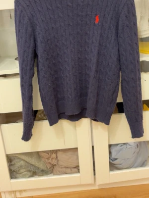 Mörkblå kabelstickad tröja Polo Ralph Lauren - Mörkblå kabelstickad tröja från Polo Ralph Lauren med klassisk röd broderad logga på bröstet. Tröjan har rund halsringning och långa ärmar, perfekt för lager-på-lager-stil. Mjuk och skön stickning som passar till många outfits. Bara att skriva vid vidare frågor