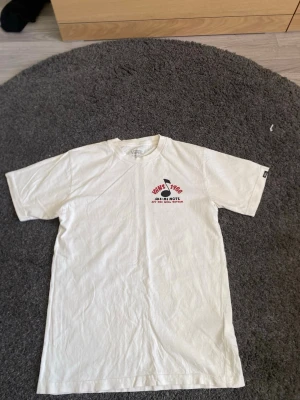 Vit Vans t-shirt med tryck - Vit t-shirt från Vans med coolt tryck fram och stort motiv på ryggen med texten 'Vans 1966' och en hund som DJ. Skön passform och klassisk rund hals. Perfekt för dig som gillar streetwear och vill sticka ut med en unik design.