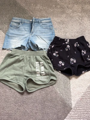 Shorts - Säljer ett 3-pack shorts: ett par ljusblå jeansshorts från H&M med rå kant, ett par svarta shorts med blommigt mönster och ett par gröna mjukisshorts från H&M med trycket 'Always Believe'. Perfekta för sommaren och chill dagar.