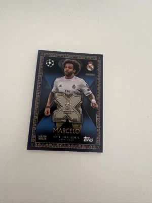 Topps UCL Decades samlarkort Marcelo - Samlarkort från Topps med Marcelo i Real Madrid, UCL Decades 2010-2020. Kortet har en stor silverfärgad X-logga och detaljer i guld och blått. Perfekt för dig som gillar fotboll och samlar på unika sportkort.                                                      Marcelo UCL DECADES  UCLD-MC Max attax extra 2025-26  TOPPSKICK - Nyss draget i ett pack och är varsamt hanterat.