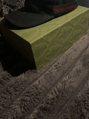 Svart keps från Gucci med röd-grön rand - Snygg svart keps från Gucci med diskret GG-mönster i tyget och en klassisk röd-grön rand på sidan. Kepsen har böjd skärm och justerbar passform. Materialet är textil och insidan har märkets etikett.