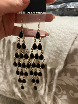 Svarta droppformade statementörhängen - Snygga och långa örhängen från Accessorize med flera svarta droppformade hängen i guldtonad metall. Perfekt för dig som gillar att sticka ut med dina accessoarer. Örhängena har en elegant och trendig design med många små svarta detaljer.